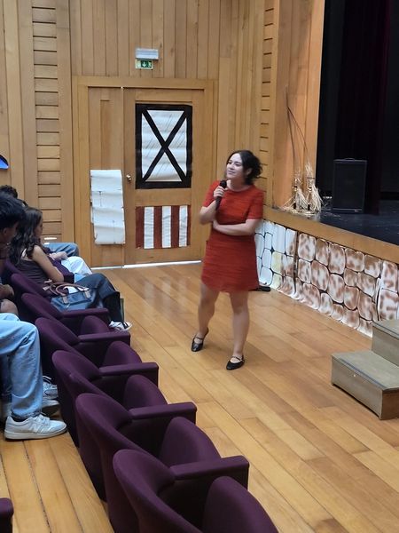 Mulher de vestido vermelho, de microfone na mão, ouve os estudantes